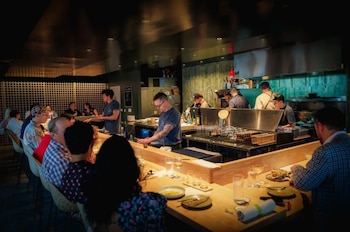 Vista interior del restaurante Anjin con clientes sentados en una larga barra de madera, frente a una cocina abierta donde varios chefs trabajan. Vasos y platos sobre la barra