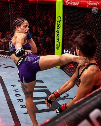 Una luchadora vestida con indumentaria morada le conecta una patada en el rostro a su oponente en un octágono de artes marciales