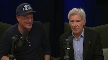 Dos hombres, Woody Harrelson y Harrison Ford, sentados frente a micrófonos; Harrelson sonríe con gorra, Ford habla con traje. Fondo oscuro.