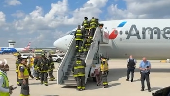 Bomberos con uniforme suben por una escalera móvil a un avión comercial en una pista junto a policías y vehículos de asistencia