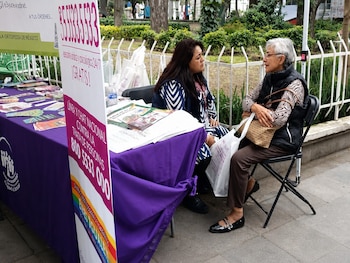 Dos mujeres conversan junto a una mesa con folletos y carteles informativos en un espacio exterior con árboles