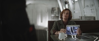 Una mujer de mediana edad con cabello rojizo y chaqueta verde oliva sostiene una tableta holográfica interactiva en un entorno interior futurista