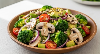 Primer plano de una ensalada en un cuenco de madera, con champiñones en rodajas, brócoli, aguacate en cubos, cebolla morada, tomates cherry y semillas.