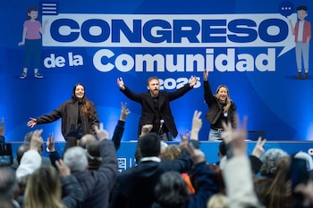Tres personas saludan desde un escenario con un cartel azul de fondo ante un público que levanta las manos