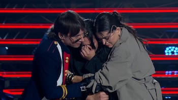 Joaquín Levinton, con traje azul y rojo, y Natalie Pérez, con chaqueta gris, abrazan a una joven que cubre su rostro con las manos en un escenario iluminado