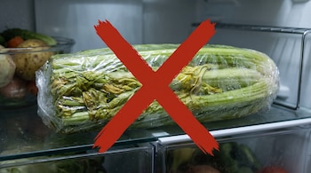 Un manojo de apio envuelto en plástico transparente sobre la repisa de un refrigerador con un símbolo de cruz roja encima.
