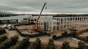 Una grúa industrial sostiene un tanque metálico junto a una estructura blanca en construcción, vehículos de carga y un edificio industrial
