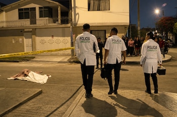 Tres agentes con batas de la Policía Criminalística caminan de noche hacia un cuerpo cubierto en el suelo delimitado por una cinta amarilla