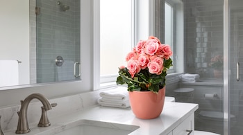 Interior de baño moderno con lavabo, grifo metálico, encimera de mármol, maceta con flores rosadas, toallas blancas, ventana y ducha con azulejos grises.