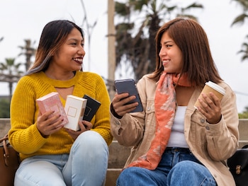 Dos mujeres sentadas al aire libre sostienen cajas de cosméticos, un teléfono móvil y un vaso de café desechable