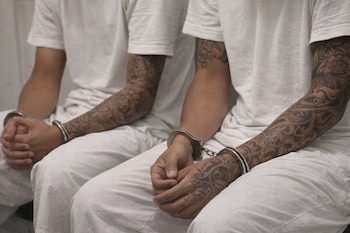 Imagen de dos personas sentadas, con uniformes blancos, esposas en sus muñecas y brazos y manos extensamente tatuados con patrones ornamentales.