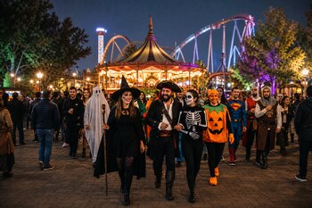 Personas vestidas con disfraces de pirata, bruja, esqueleto y calabaza caminan de noche frente a un carrusel iluminado y una montaña rusa.