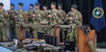 Fusiles de color arena sobre soportes de madera y cargadores en primer plano con soldados en uniforme de camuflaje al fondo