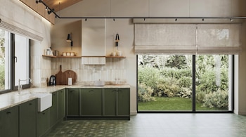 Cocina interior con gabinetes verdes, encimeras claras, fregadero, grifo, tetera, tablas de cortar, estantes, ventanal grande con vista a un jardín, piso de baldosas.