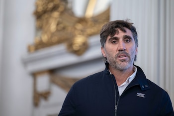 Retrato de un hombre de mediana edad con barba, cabello oscuro despeinado, y chaqueta azul marino con un logo, frente a un fondo borroso de adornos dorados