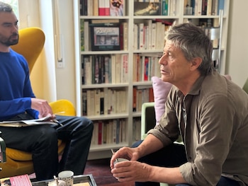 Emmanuel Carrère, de mediana edad y cabello entrecano, sentado en un sofá verde sosteniendo una taza, frente a un entrevistador joven con papeles, siendo grabado por un smartphone