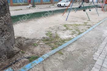 Área de tierra con un árbol, un cordón azul, una acera de hormigón y una calzada con un coche blanco al fondo, con columpios visibles