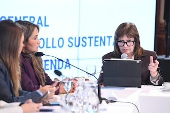 Mujer con gafas y chaqueta marrón habla en un micrófono, con una tablet y otras dos mujeres en una mesa. Pantalla de fondo con texto azul