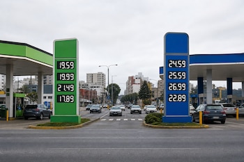 Dos letreros de estaciones de servicio, uno verde y uno azul, muestran precios digitales a los lados de una avenida con tráfico.