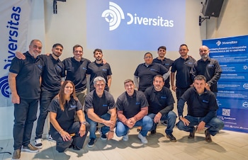 Trece personas, doce hombres y una mujer, con polos negros y logo, posan. Detrás, una pared con el logo 'Diversitas' proyectado, un banner y una bandera