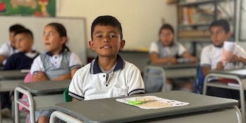 Un niño sentado frente a un pupitre con un dibujo y unas tijeras, junto a otros compañeros en un aula escolar