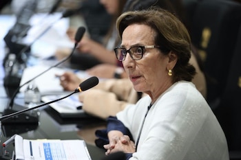 Mujer con gafas y prenda blanca habla ante un micrófono en una mesa de reuniones rodeada de carpetas y personas