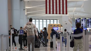 Vista interior de una terminal de aeropuerto con pasajeros y equipaje. Una gran bandera estadounidense cuelga del techo. Paneles de información de vuelos visibles al fondo