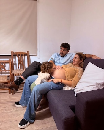 Marcela Pagano sentada en un sofá con su pareja, mientras un niño pequeño se inclina para besar su vientre de embarazo expuesto