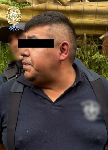 Un hombre con los ojos cubiertos por una barra negra viste una camisa oscura junto a un policía y un emblema institucional