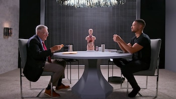 Dos hombres sentados en una mesa, con un modelo anatómico de torso, vasos, un libro, una carpeta y un frasco de pastillas