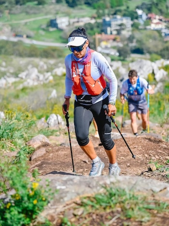 Una mujer con visera, gafas de sol y bastones asciende por un camino de tierra, seguida a la distancia por otro corredor