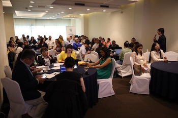 Salón de conferencias con mesas redondas y sillas blancas. Numerosas personas sentadas. Un hombre de pie al frente con micrófono. Laptops, teléfonos y documentos en las mesas. Banner
