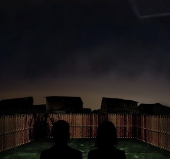 Dos siluetas de personas observan una cerca de madera, un césped y casas contra un cielo nocturno oscuro con estrellas y una luz ovalada
