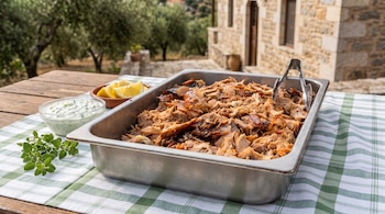 Una bandeja de metal con carne de cerdo desmenuzada sobre una mesa con mantel a cuadros, junto a salsa blanca, limones y orégano.