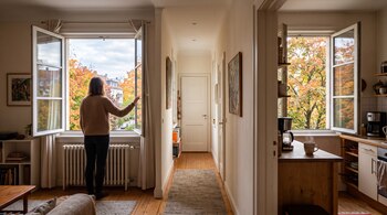 Una mujer de espaldas junto a una ventana abierta en un salón, un pasillo central y una cocina con otra ventana abierta que da a árboles otoñales.