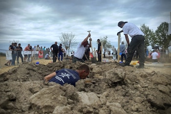 Varias personas con palas excavan tierra en un terreno mientras otros espectadores observan detrás de una valla metálica