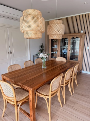 Un comedor con mesa de madera, ocho sillas de rejilla y madera clara, dos lámparas colgantes de bambú y un armario de madera detrás. Un jarrón con flores blancas sobre la mesa