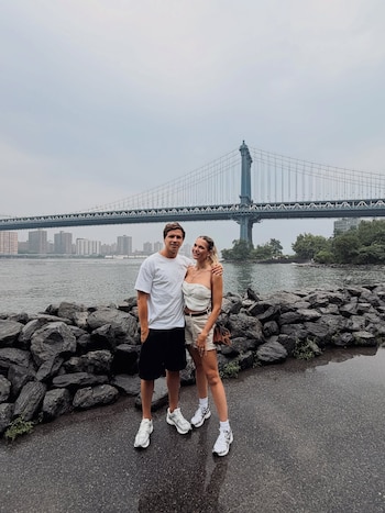 Un hombre y una mujer de pie abrazados en una orilla rocosa, con un río y un gran puente de suspensión con edificios al fondo y cielo nublado
