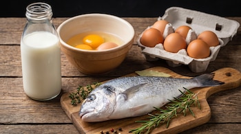 Botella de leche, un cuenco con tres huevos sin cáscara, un cartón con seis huevos, un pescado fresco, romero y tomillo en una tabla de cortar de madera.