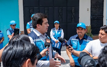Un hombre con chaleco azul habla rodeado de micrófonos y reporteros en una calle junto a personal institucional con gorras