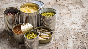 Seis latas de conserva, algunas abiertas, con frijoles rojos, trozos de piña, guisantes, frijoles en salsa y sardinas sobre una superficie.