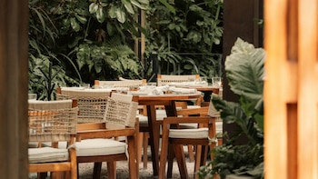 Mesas redondas de madera y sillas con cojines blancos en un patio exterior, rodeadas de abundante vegetación verde en el restaurante Esca