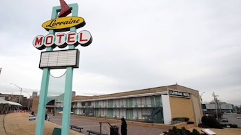 Un letrero alto con la palabra 'Lorraine Motel' en amarillo y rojo sobre un poste turquesa, con un edificio de motel de dos pisos detrás bajo un cielo nublado