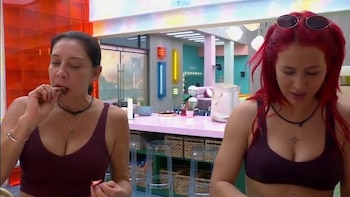 Dos mujeres, una de cabello oscuro comiendo fruta y otra de cabello rojo con gafas de sol sobre la cabeza. Un mostrador rosa y un fondo con luces de neón