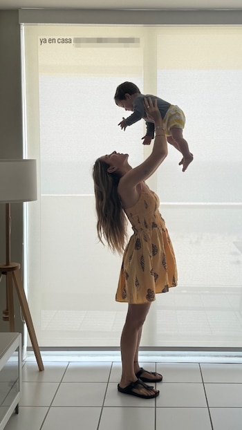 Jimena Barón, de pie y de perfil, levanta a su hijo Arturo sobre su cabeza frente a una ventana con cortinas claras