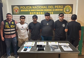 Un agente policial y cuatro hombres parados detrás de una mesa con teléfonos móviles, armas de fuego y municiones frente a un panel oficial