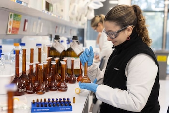 Una mujer con gafas de seguridad y guantes sostiene un matraz de vidrio ámbar junto a frascos químicos sobre una mesa de laboratorio