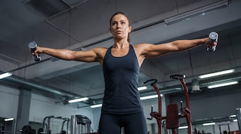 Una mujer atlética, con ropa deportiva oscura y sudor en la piel, realiza elevaciones laterales con mancuernas plateadas en un gimnasio bien iluminado.