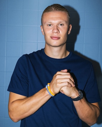 Hombre rubio con camiseta azul marino, manos entrelazadas con pulseras trenzadas y un reloj negro, frente a una pared de azulejos azules
