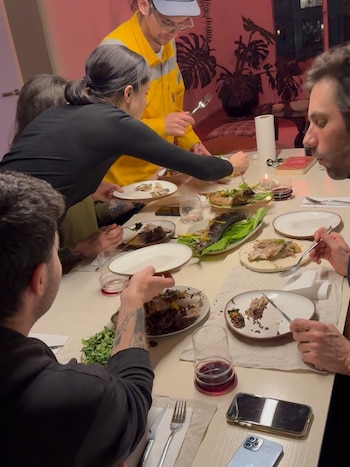 Varias personas cenan alrededor de una mesa. Hay platos con alimentos, vasos, cubiertos y teléfonos celulares. Una mujer y un hombre sirven comida en los platos